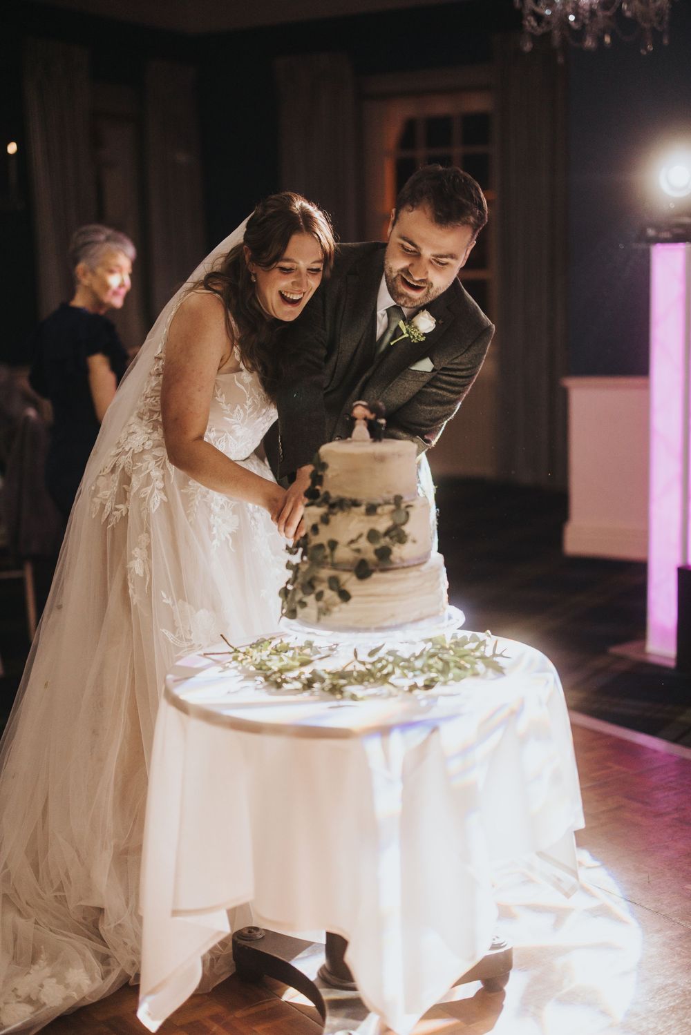 A wedding couple joyfully cuts into their illuminated white tiered cake during their evening reception ceremony.