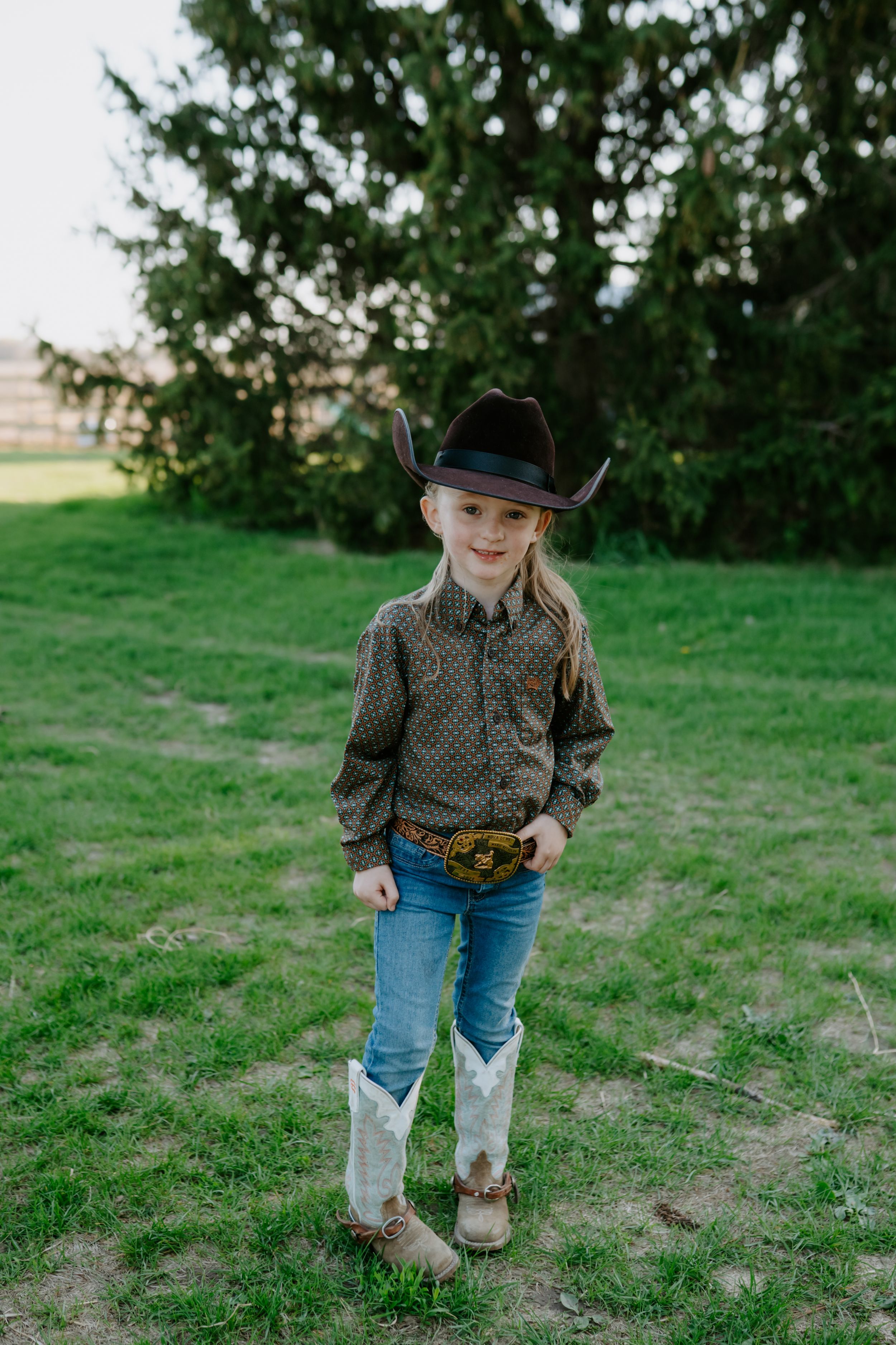 Child in western attire walking through a grassy field wearing cowboy boots and hat.