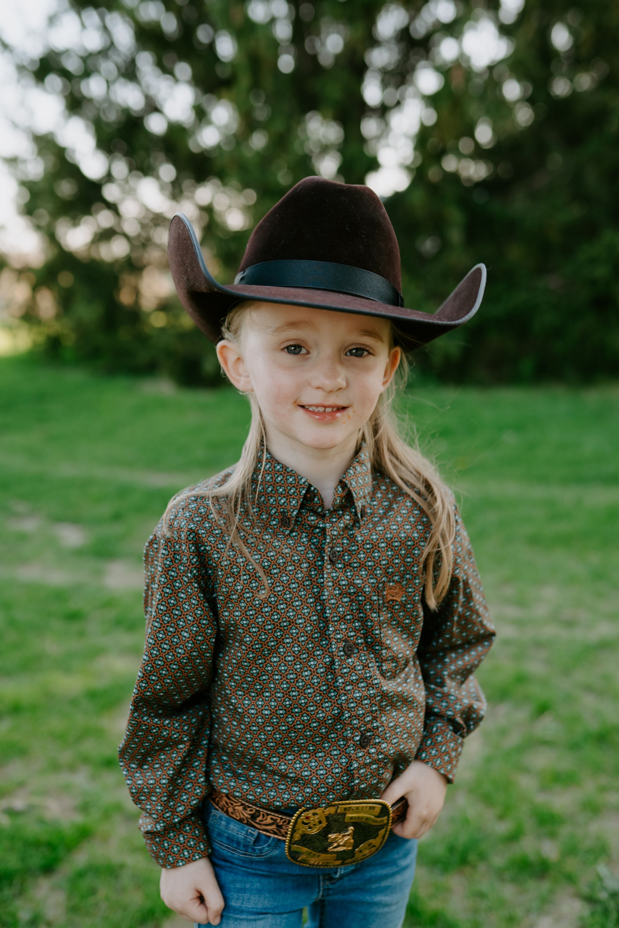 Young person in western wear and cowboy hat posing in a green field.