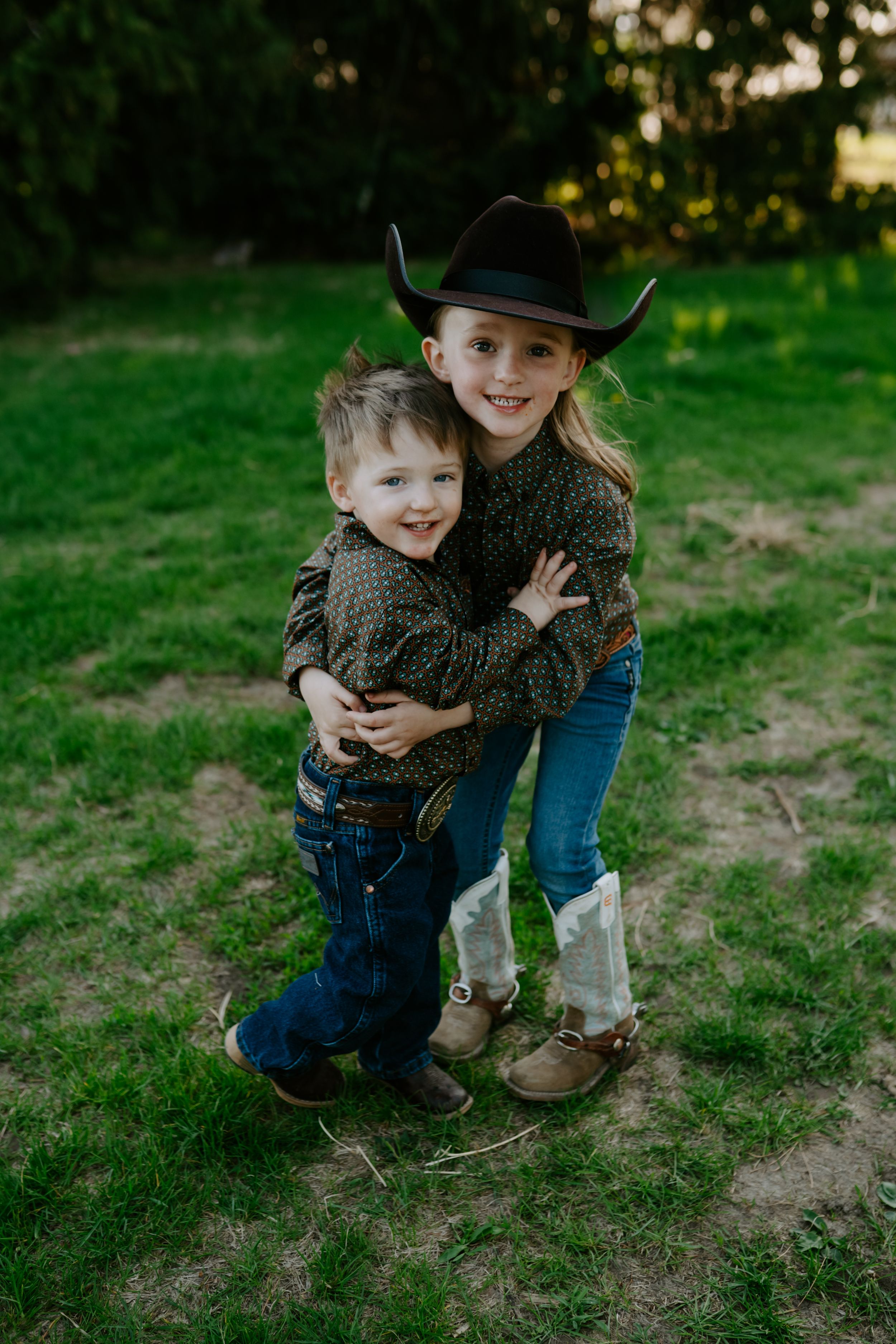 Two children in western wear share a sweet embrace while standing on grass in natural lighting.