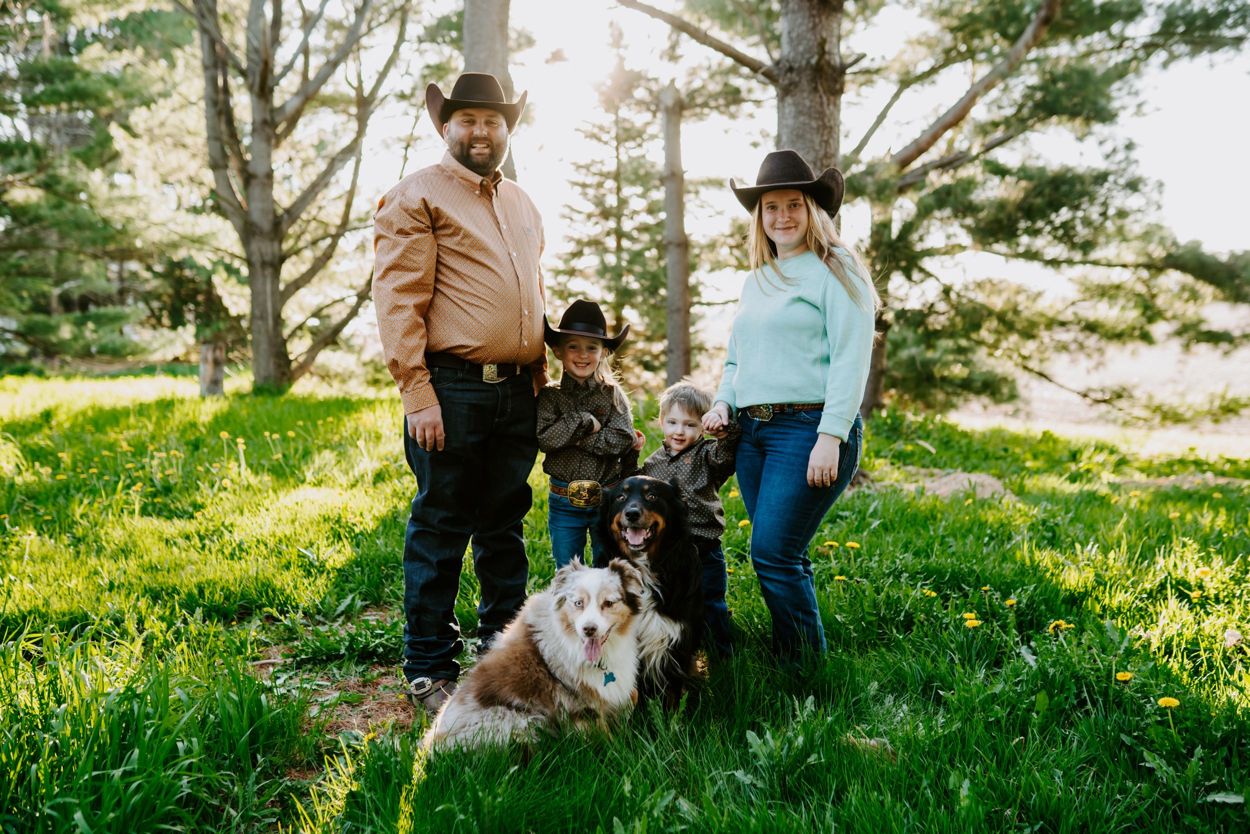 A family poses with their dog in a sunlit forest clearing wearing western-style clothing.