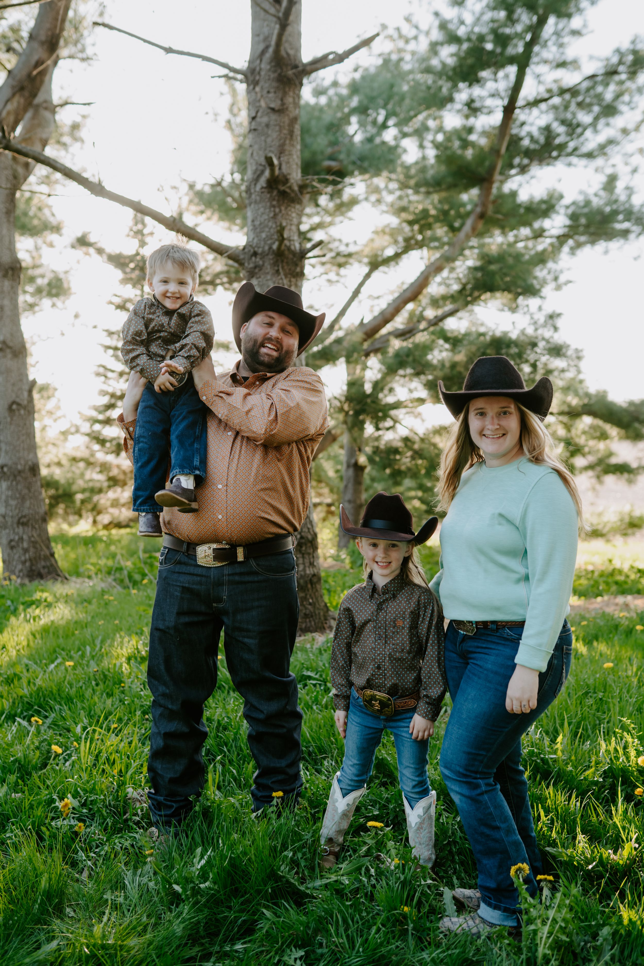 A family in western attire poses together among pine trees on a grassy hillside.