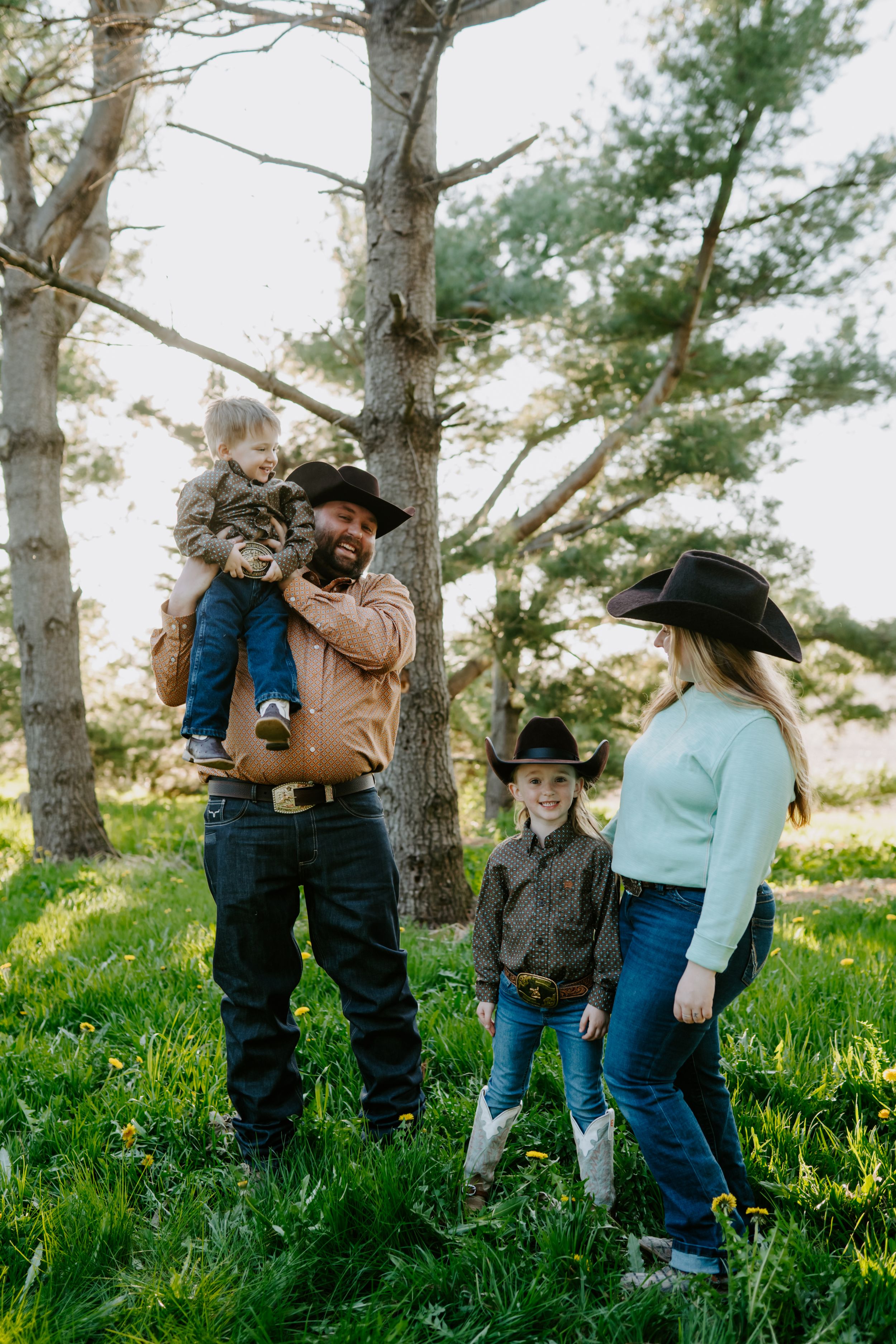 A family in western wear stands together among tall pine trees in afternoon sunlight.