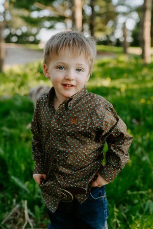 A young child in a patterned button-up shirt smiles while standing in green grass.