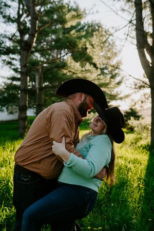 A romantic western-themed couple embrace in a sunlit grassy field wearing cowboy hats.