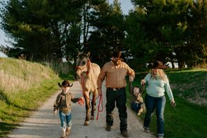A family in western attire walks down a dirt path with their horse on a sunny day.