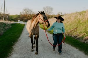 A horse and person in western attire walk together on a gravel path through a scenic countryside setting.