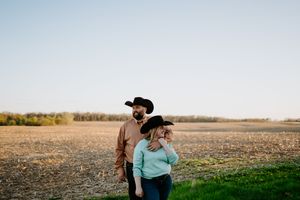 Two people in western wear stand together in an open field at sunset.