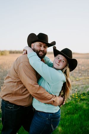 A couple in cowboy hats share an embrace in a field during golden hour.