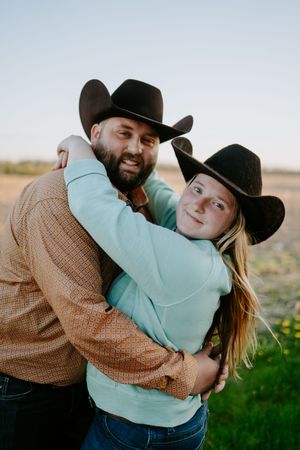 A couple in cowboy hats and western attire embrace while smiling at the camera.