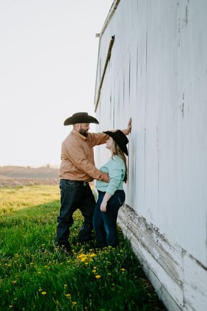 A couple in western wear share an intimate moment against a white barn wall surrounded by spring grass.