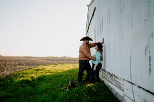 A couple in western wear share a romantic moment against a white barn wall at sunset.
