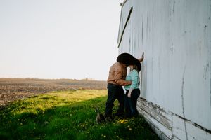 A couple in western attire share a tender moment against a white barn wall at sunset.