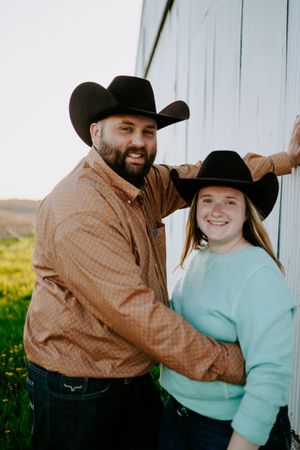 A couple in cowboy hats pose together against a white barn wall in a rural setting.