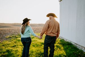 A parent and child walk together through a grassy field at sunset beside a white wall.