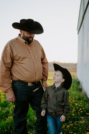 A cowboy-hatted pair stand together in a wildflower field beside a white building.