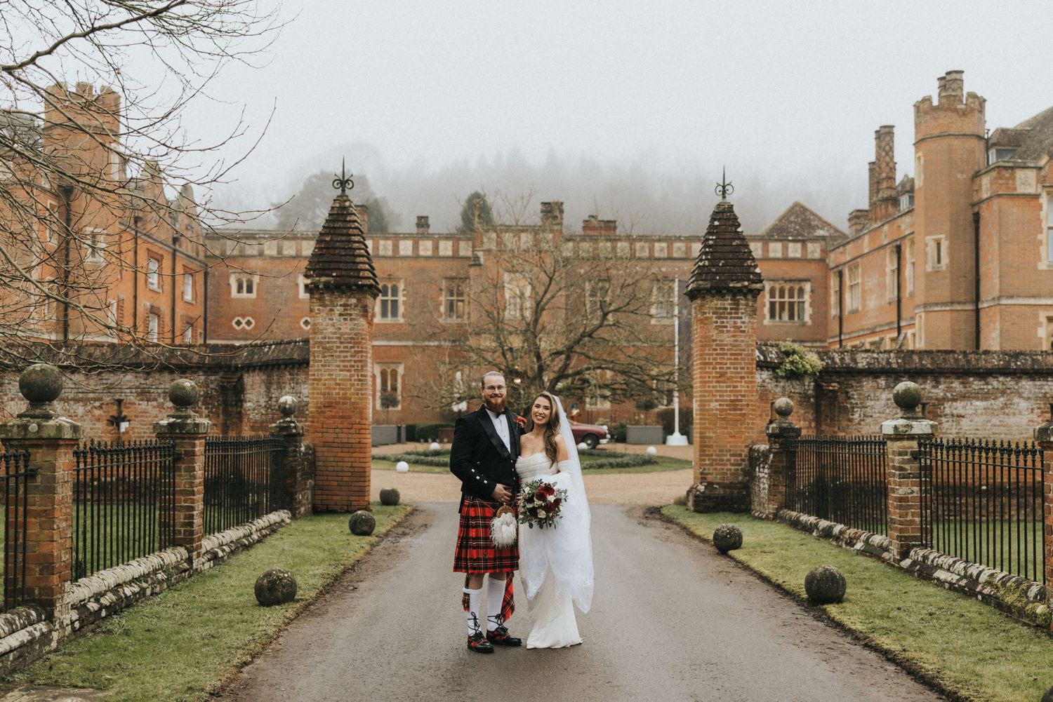 Winter wedding couple walks along a historic brick pathway flanked by grass and buildings in a courtyard setting.