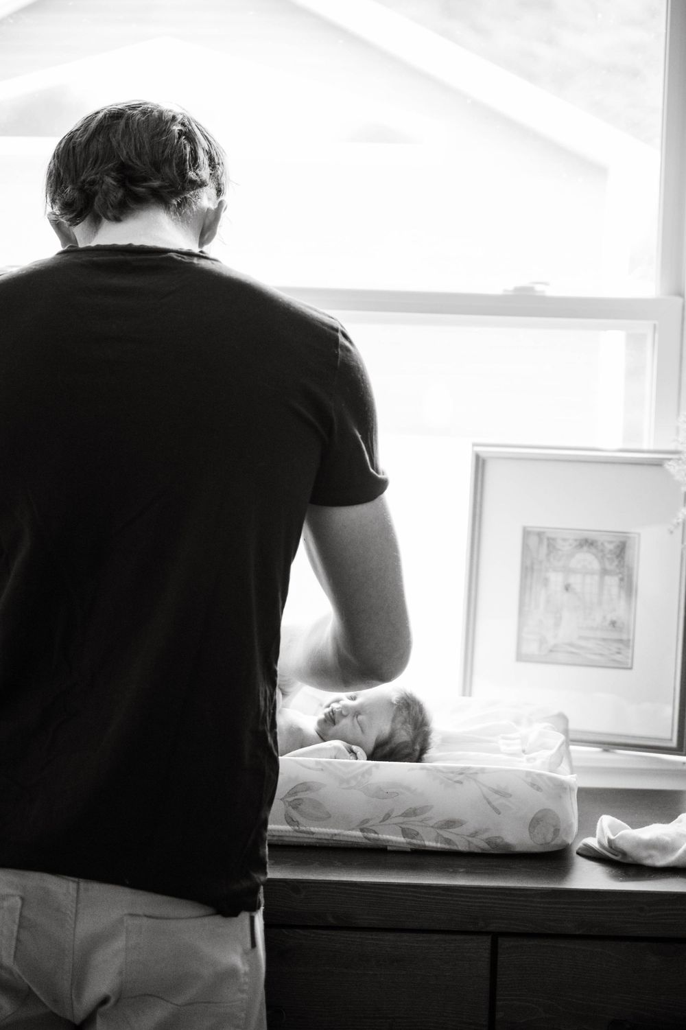 A parent leans over a changing table beside a bright window while tending to their infant.
