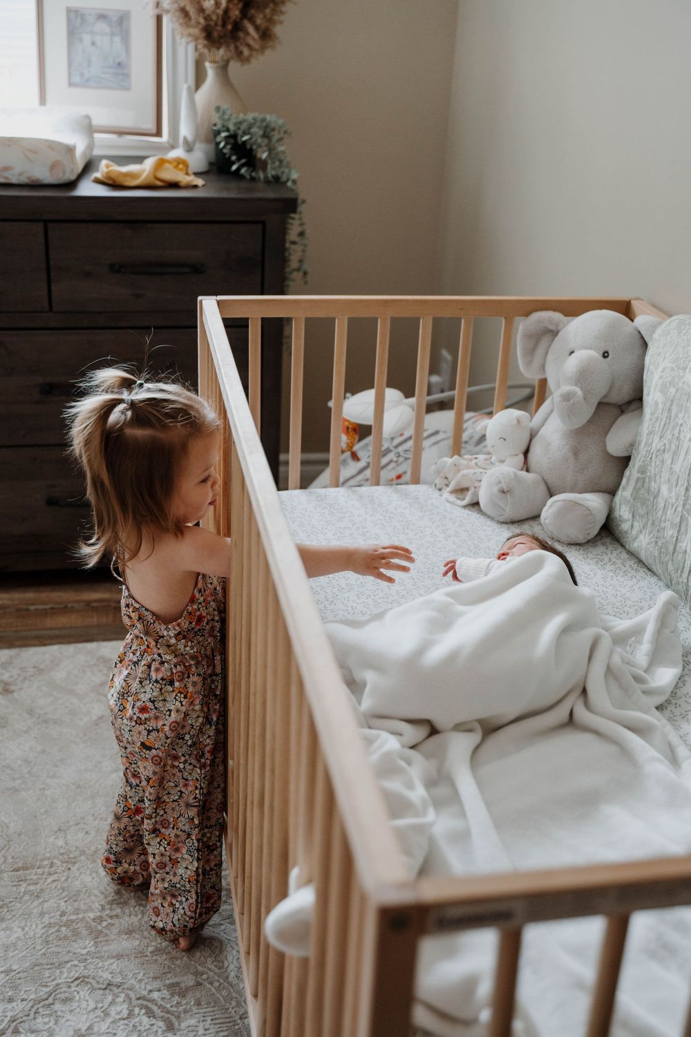 A toddler in a floral dress reaches into a wooden crib with stuffed animals and white bedding inside.
