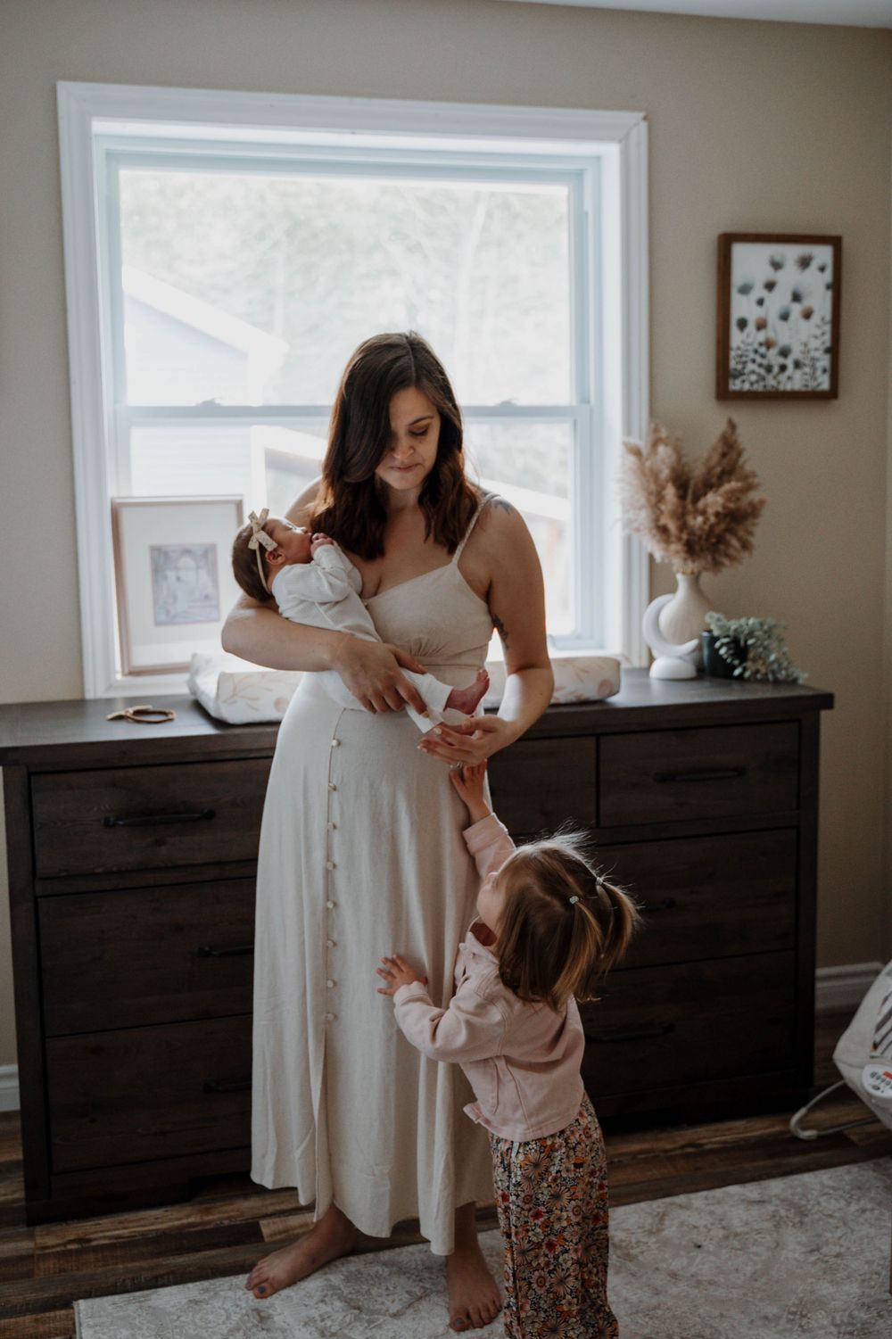 A tender family moment in front of a bright window with a white dress and toddler reaching up.