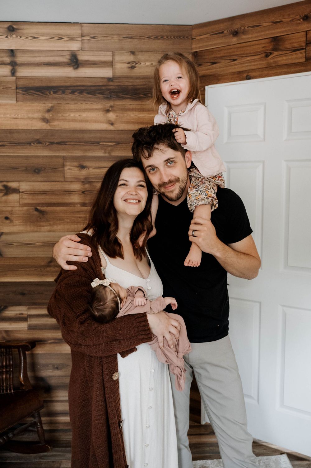 A joyful family photo with wooden wall backdrop shows parents holding their toddler who sits on father's shoulders.