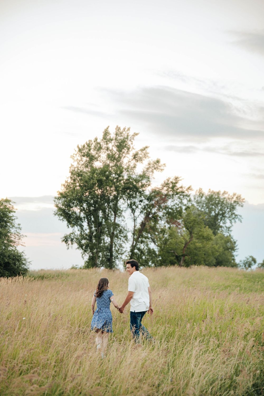 A couple walking hand in hand through a serene meadow with tall grass and trees in the background.