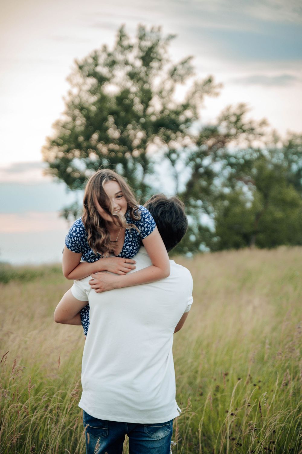 Couple embraces in a field of tall grass during sunset with clouds in the background.