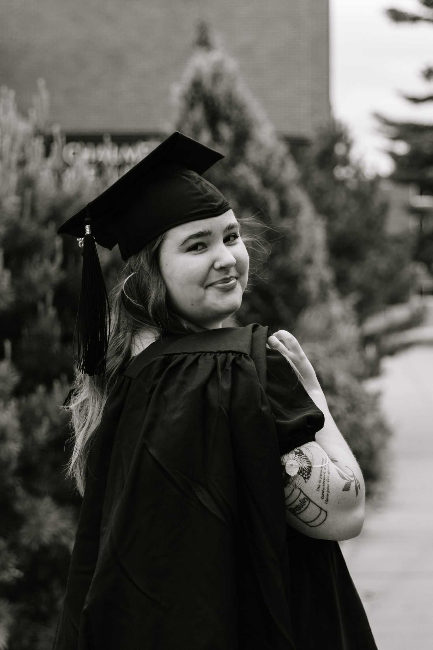 Graduate in black cap and gown poses joyfully by tree trunk during outdoor graduation photo session.