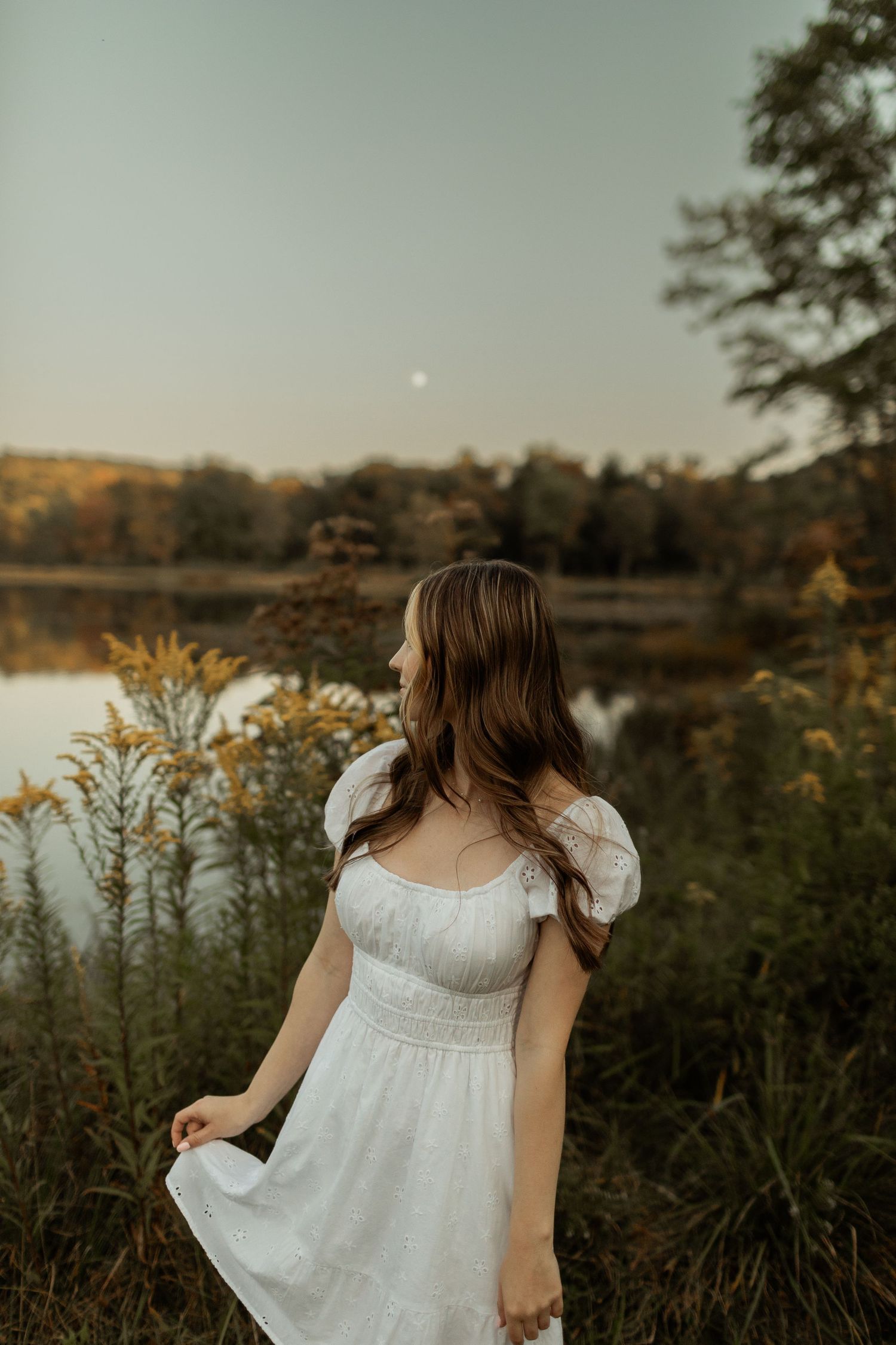 Family gathered on sandy lakeshore during misty summer evening in white dresses against wooded background.