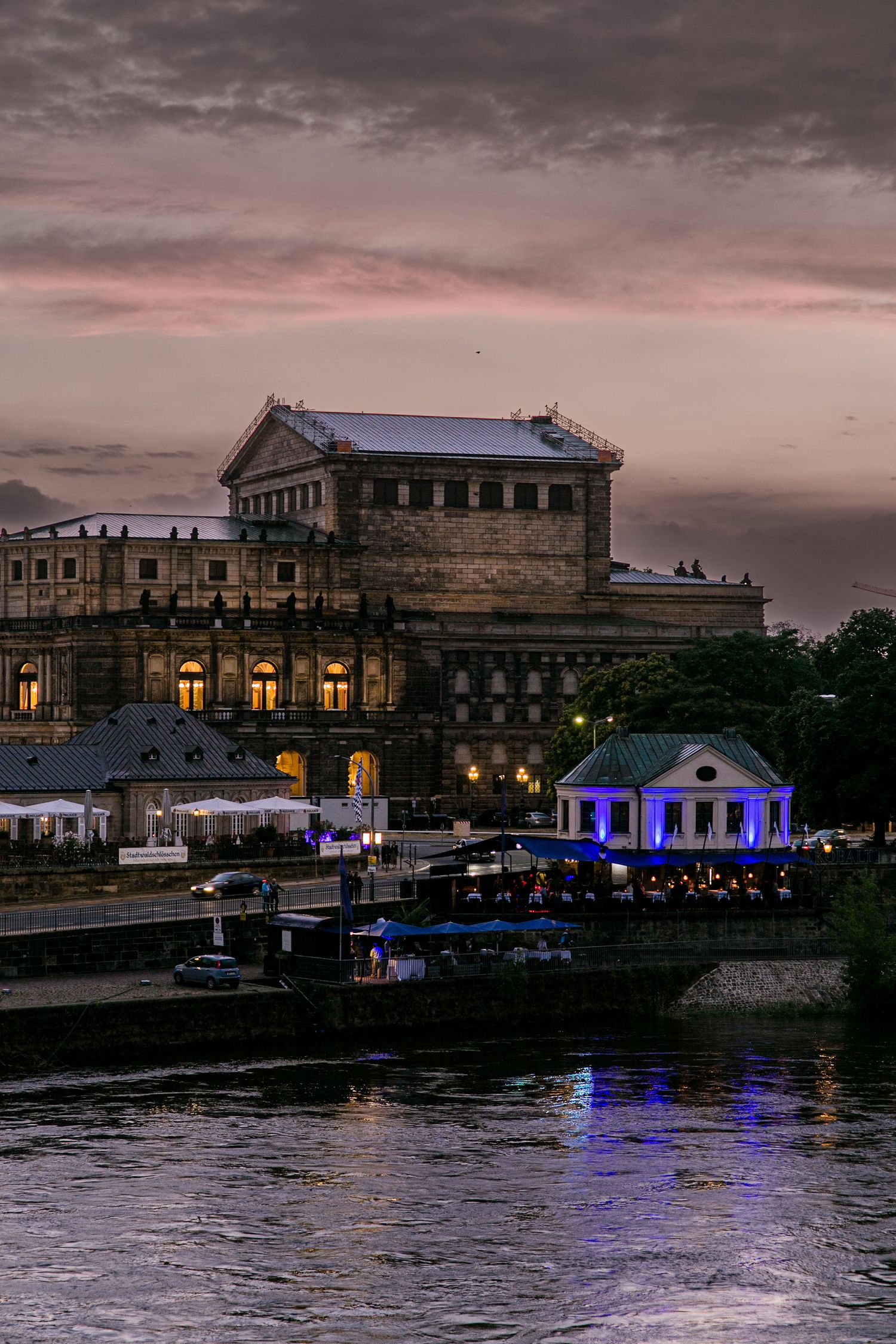 Panoramic view of illuminated buildings along a riverfront at dusk with blue lighting reflected on dark waters.
