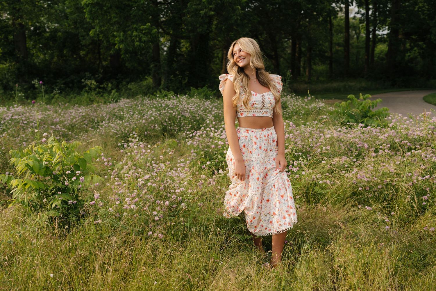 A person in a floral summer dress stands among tall grass and wildflowers in a sunlit meadow near a wooded path.