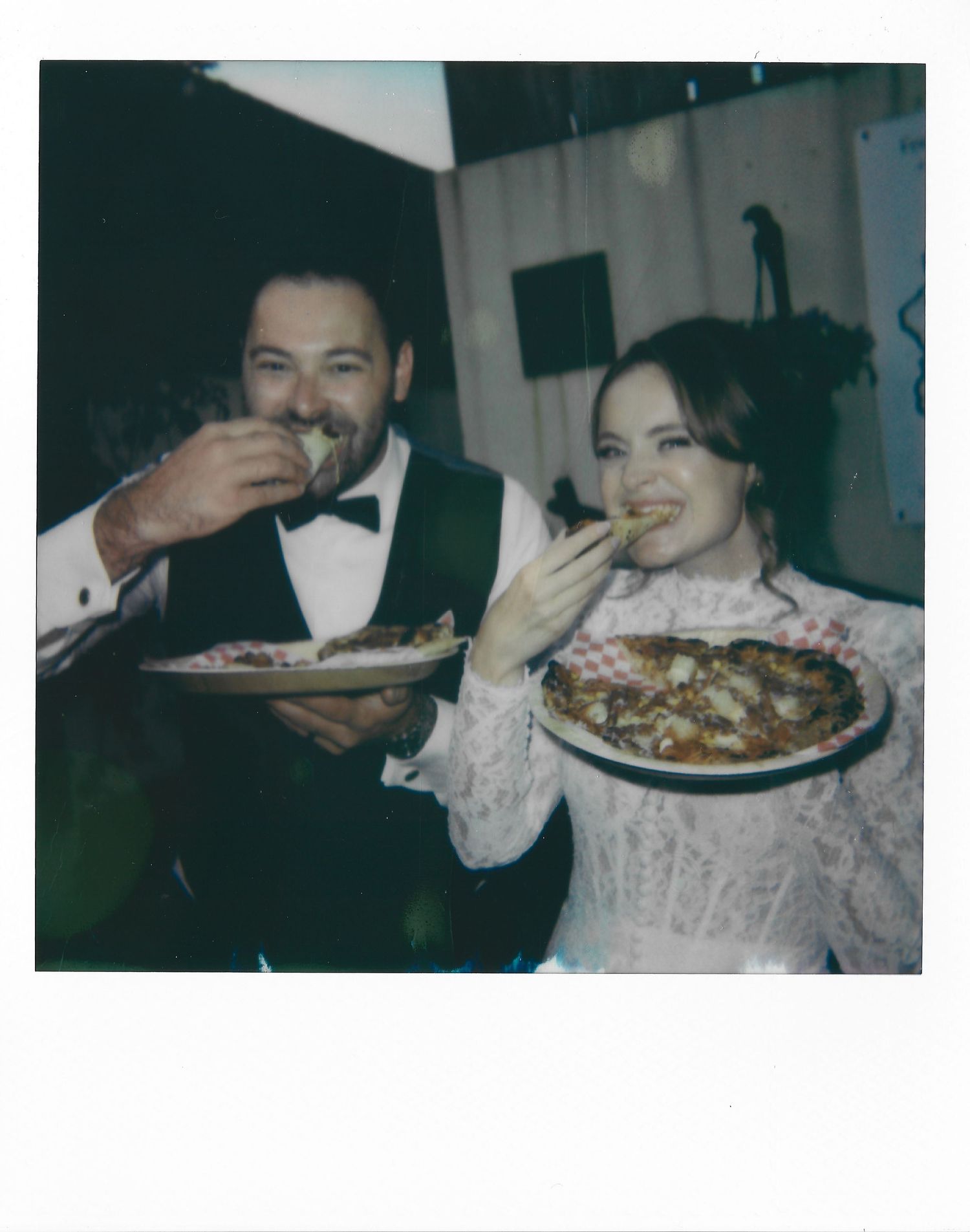 Elegant couple in formal attire share a laugh while enjoying plates of food at what appears to be a wedding reception.