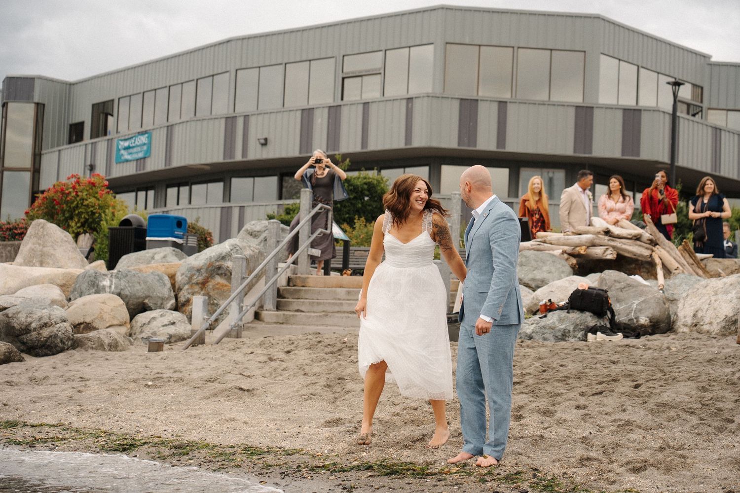 A couple celebrates their wedding day on a rocky beach near a large commercial building.