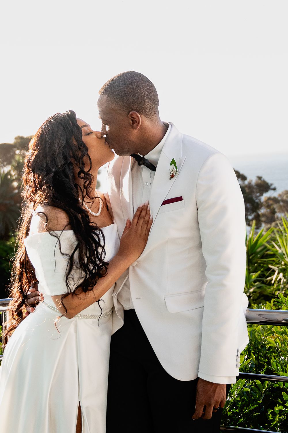 A romantic couple shares an intimate kiss wearing white formal attire against a scenic outdoor backdrop.