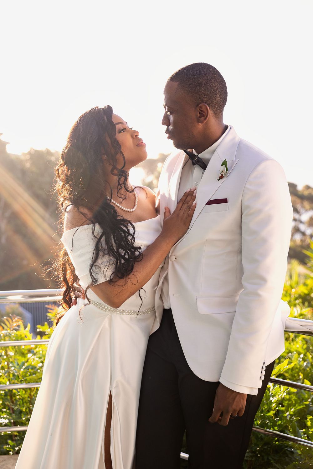 A romantic couple in formal white attire share an intimate moment at sunset on a balcony with glowing golden light.