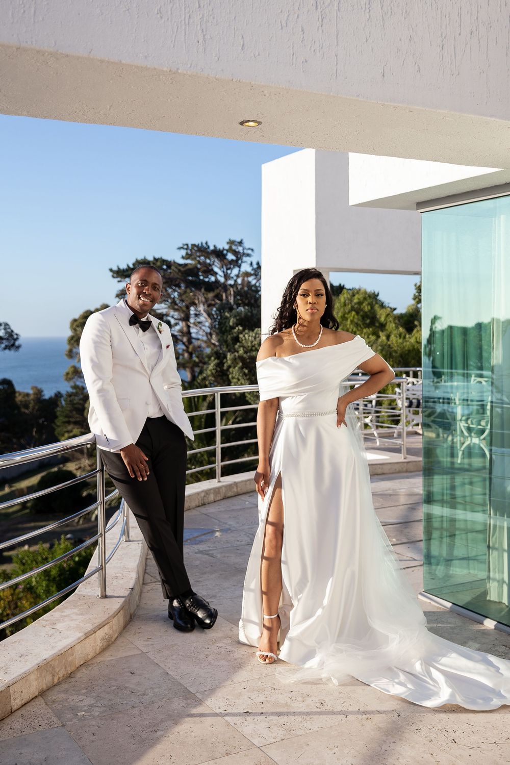 A couple poses on a modern balcony overlooking the ocean, with the bride wearing an elegant white gown with a high slit.