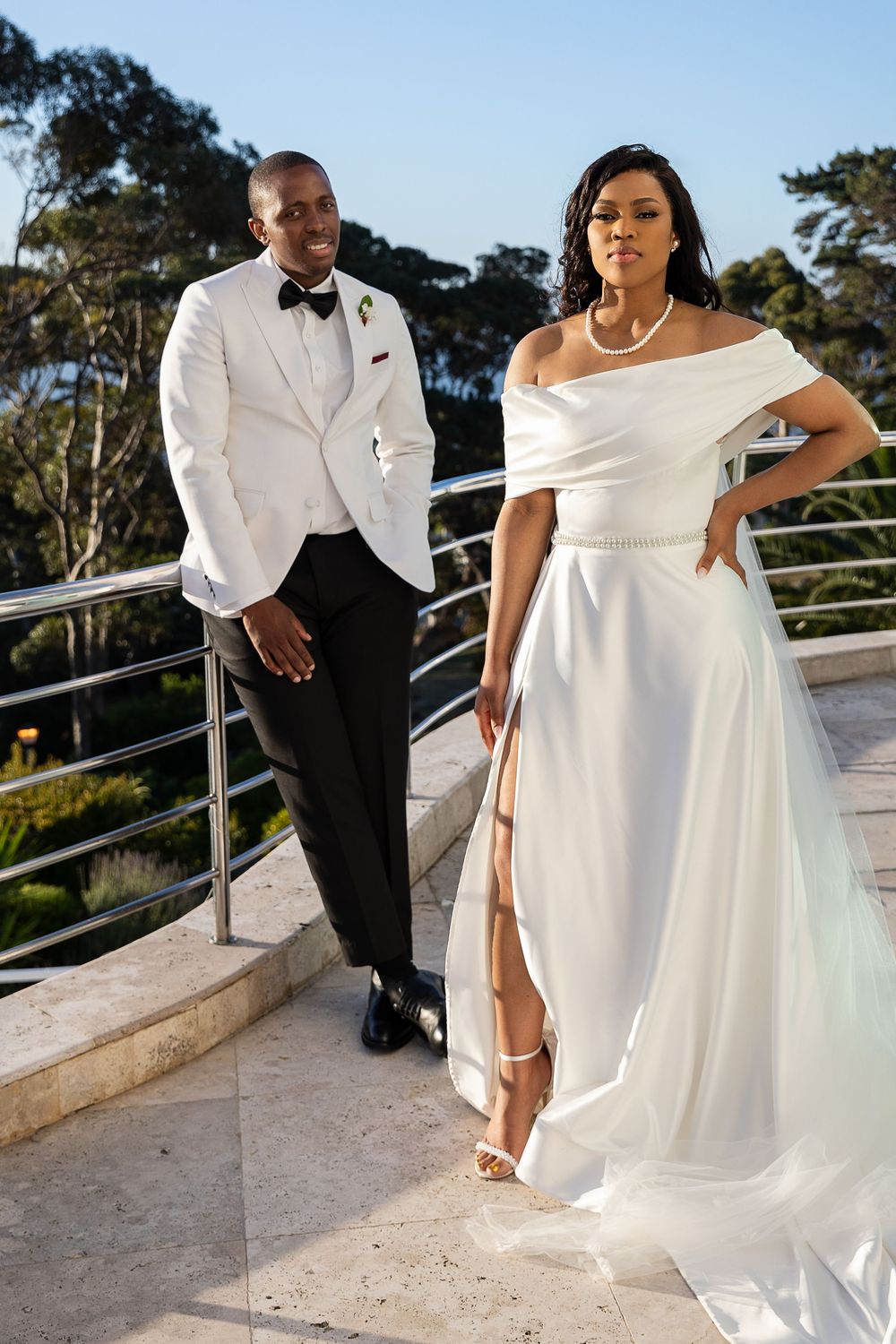 Elegant couple poses on outdoor terrace, wearing formal white wedding attire with scenic background.