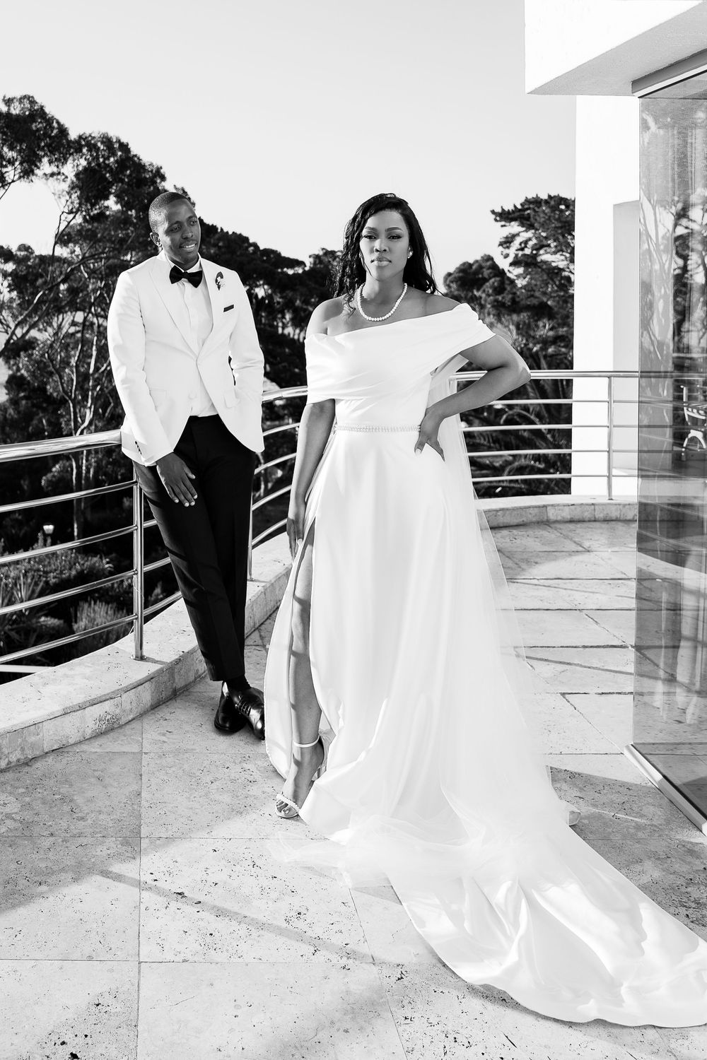 A couple stands on a balcony during their wedding, with the groom in a white tuxedo and the bride in an elegant white gown.