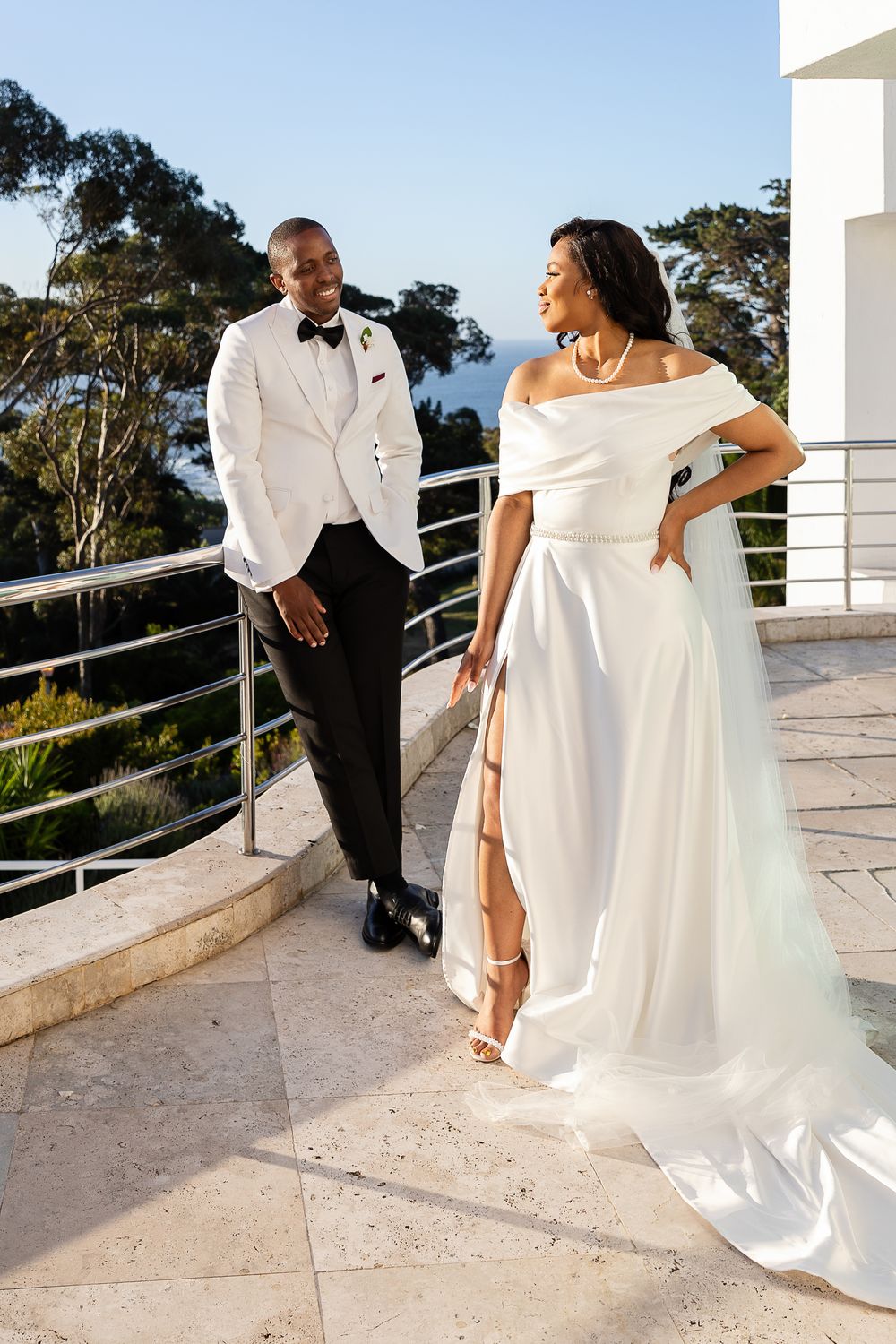 Elegant wedding couple in white dress and black tuxedo pose on outdoor terrace during golden hour.