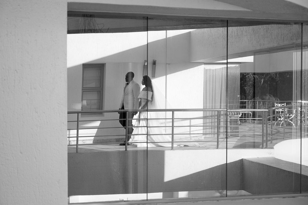 Two people stand in a sleek modern architectural space with strong diagonal shadows on white walls and glass panels.