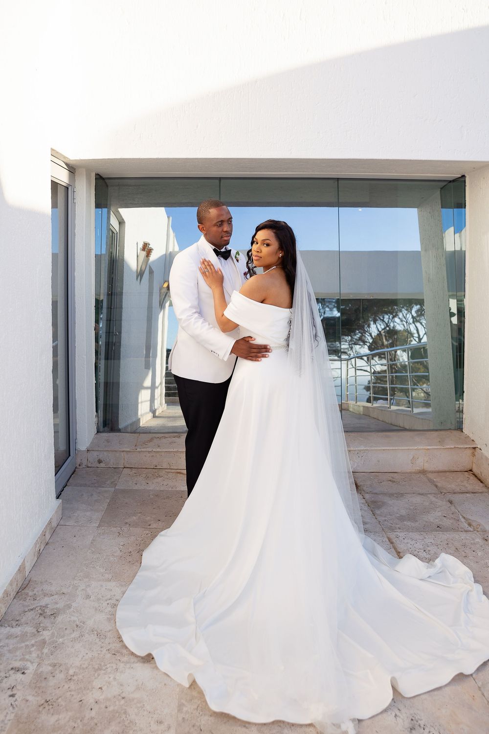 A bride in a flowing white gown and veil stands with her groom in a white tuxedo jacket on a modern stone balcony.