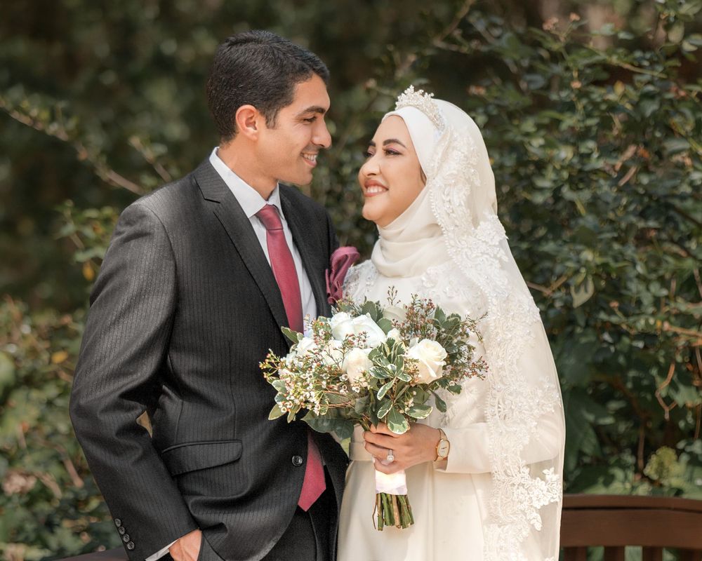 Muslim bride in white hijab and lace gown holds bouquet beside groom in gray suit at outdoor wedding.