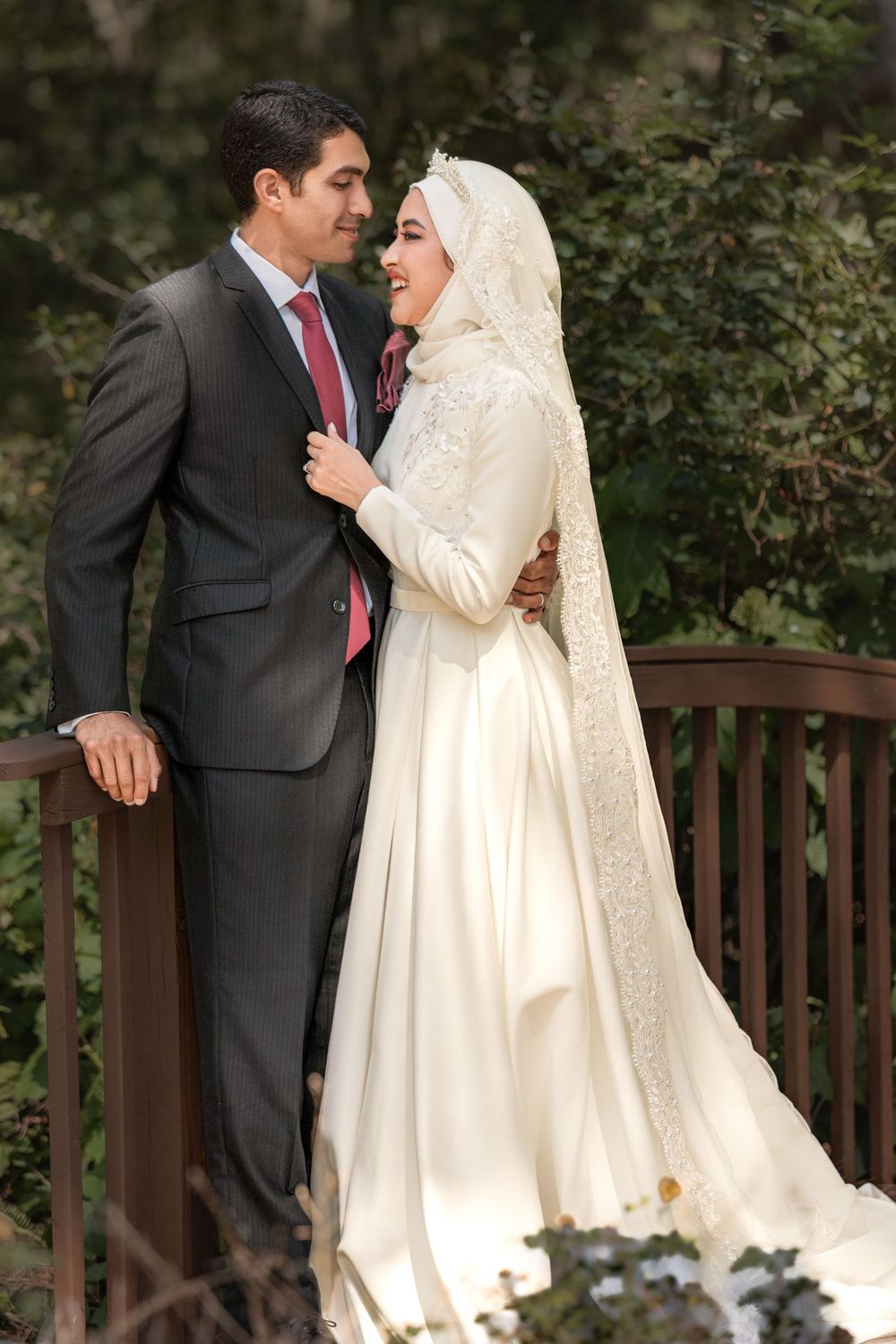 Muslim bride and groom in wedding attire pose on a wooden bridge surrounded by lush greenery.