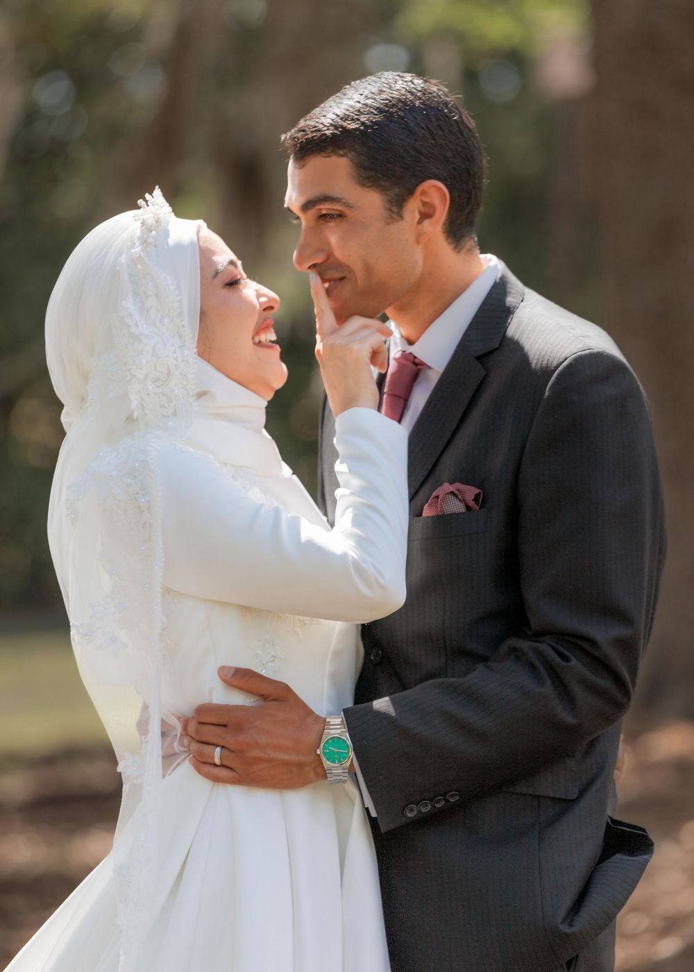 Happy couple in wedding attire sharing an intimate moment outdoors, with bride in white hijab and groom in dark suit.