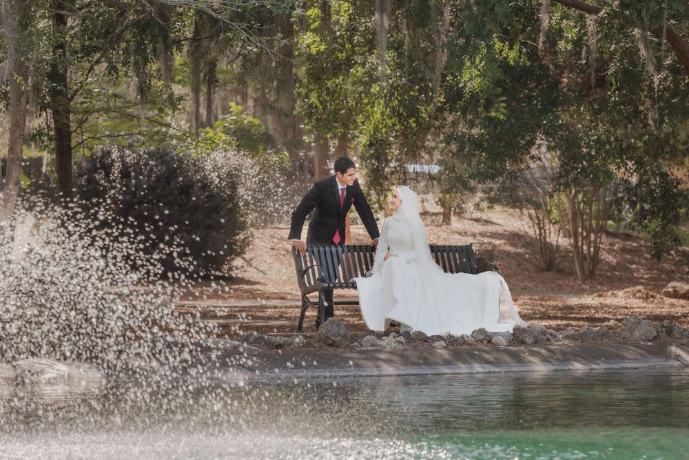 Couple sits on wooden bench by serene lake with water splashing in foreground surrounded by trees.