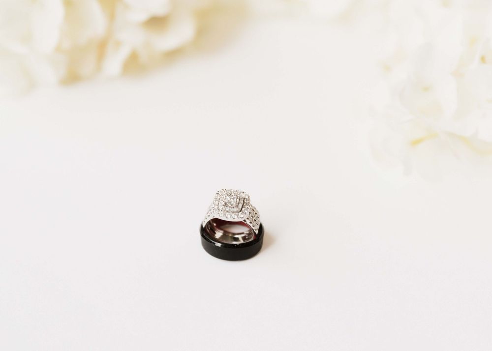 A diamond engagement ring and black wedding band sit together on a white textured surface.