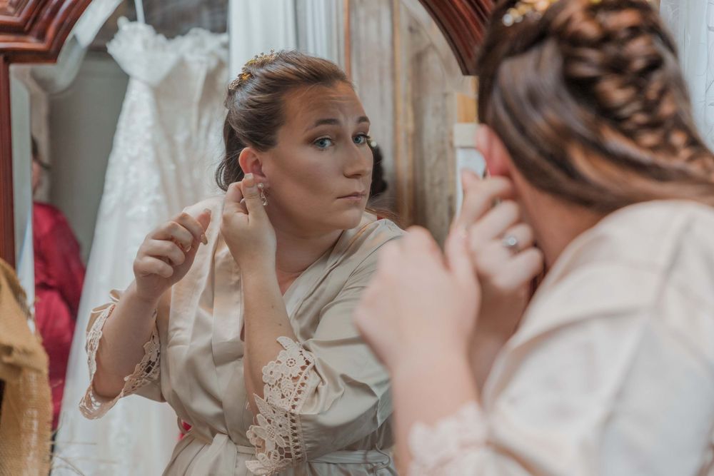 Someone in a cream colored outfit applies makeup while looking in a mirror in what appears to be a dressing room.