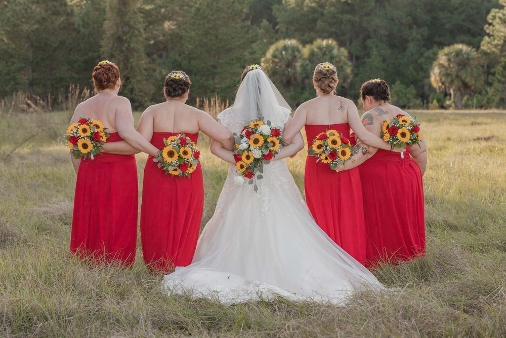 A bridal party in bright red bridesmaid dresses and white wedding gown pose together in a sunny outdoor field setting.