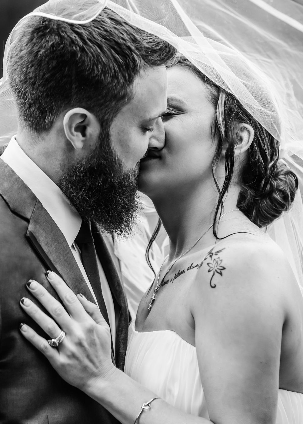 Intimate black and white wedding photo of a bearded groom in suit and bride in white dress sharing a romantic kiss.