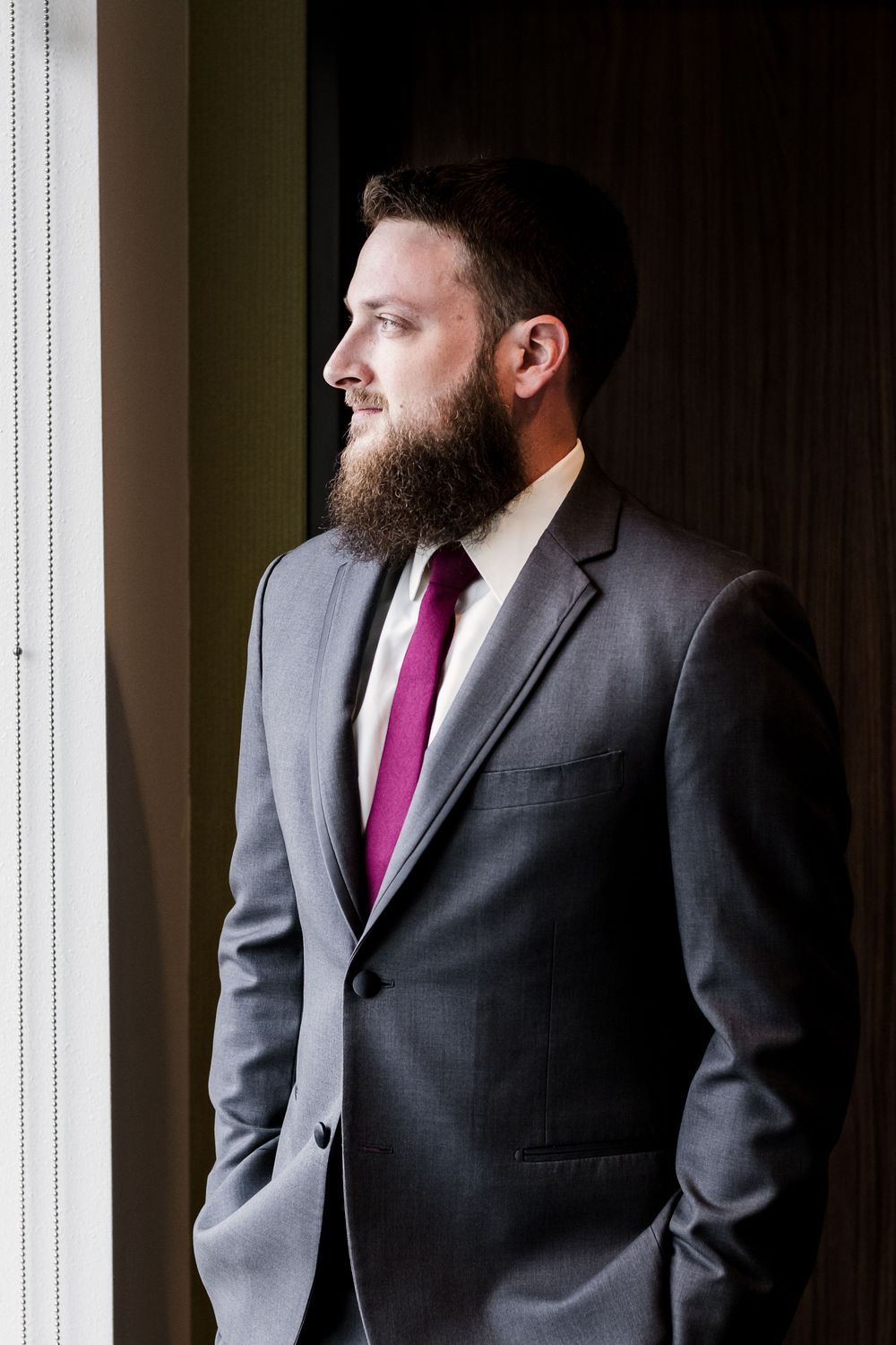 Professional portrait of person in gray suit with burgundy tie looking contemplatively out window in dramatic lighting.